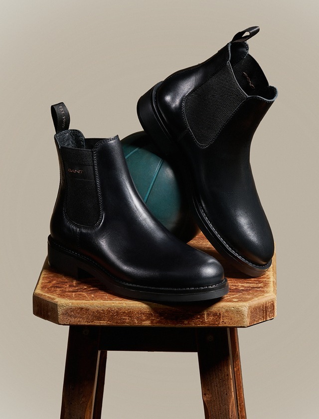 Schwarze Lederstiefeletten auf einem Holzschemel, mit einem blauen Ball im Hintergrund.
