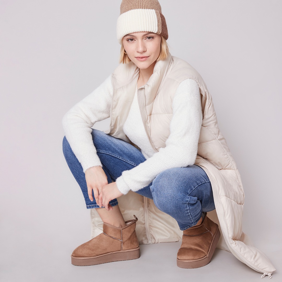 Eine Person kniet in modischer Winterkleidung, bestehend aus einem weißen Pullover, einer hellen Weste und einer Beanie. Sie trägt Jeans und braune Stiefel.