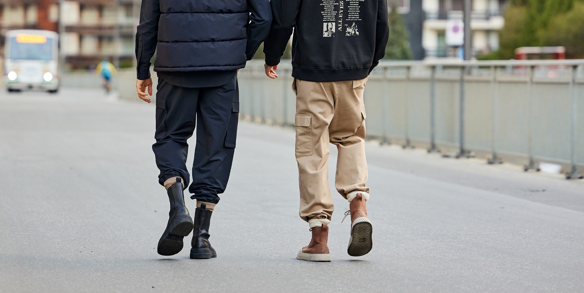 Zwei Personen gehen Hand in Hand auf einer Straße, eine trägt einen schwarzen Hoodie und beige Hosen, die andere ein dunkelblaues Outfit mit einer gesteppte Weste.