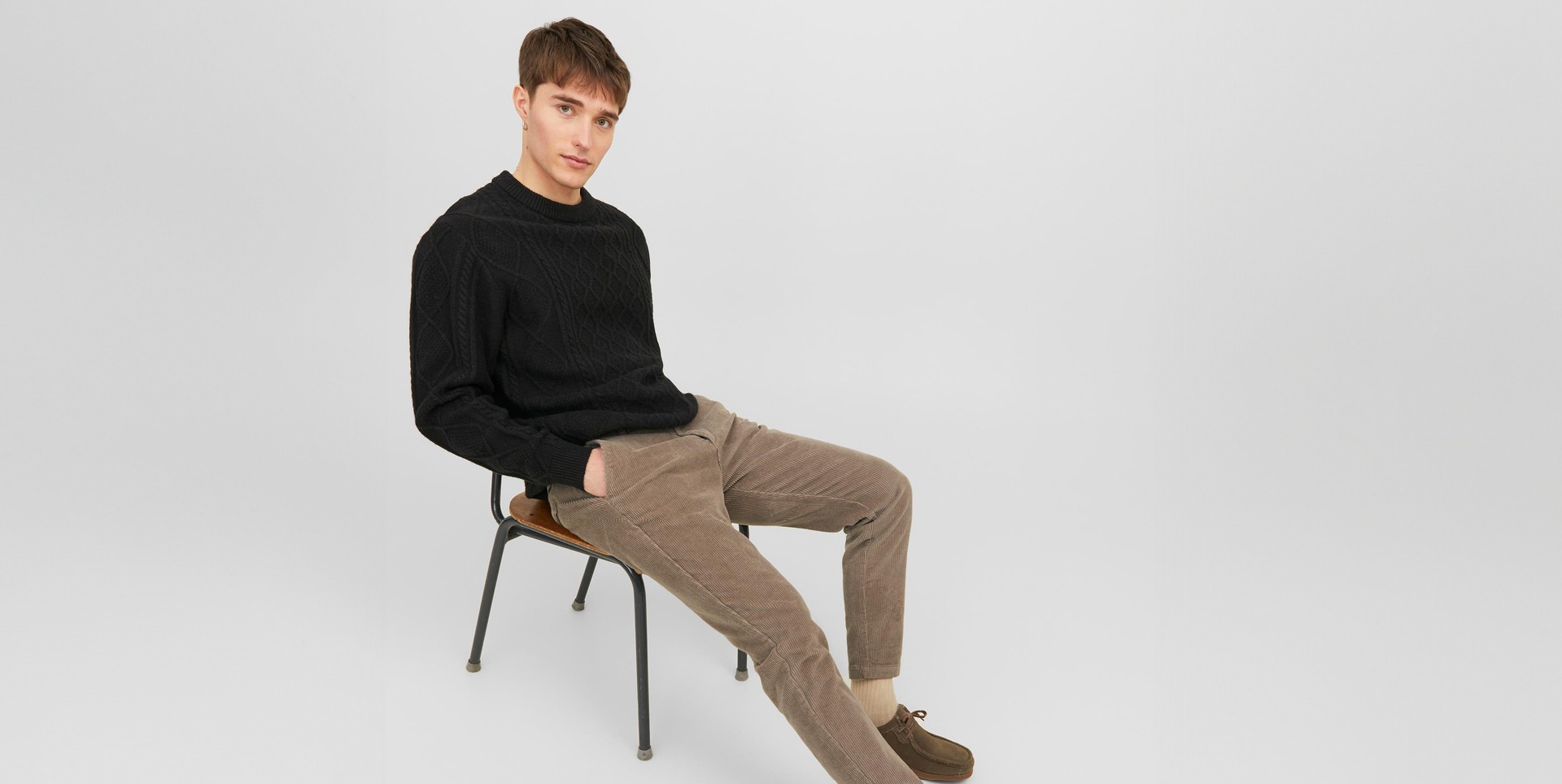 Ein junger Mann sitzt auf einem Stuhl, trägt einen schwarzen Pullover und braune Cordhosen. Ein junger Mann sitzt auf einem Stuhl, trägt einen schwarzen Pullover und braune Cordhosen.