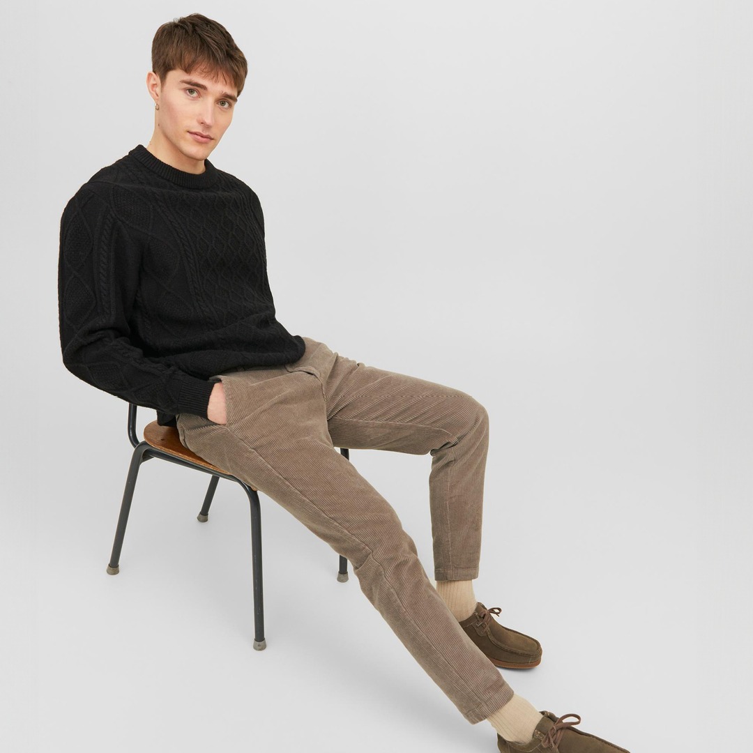 Ein junger Mann sitzt auf einem Stuhl, trägt einen schwarzen Pullover und braune Cordhosen. Ein junger Mann sitzt auf einem Stuhl, trägt einen schwarzen Pullover und braune Cordhosen.