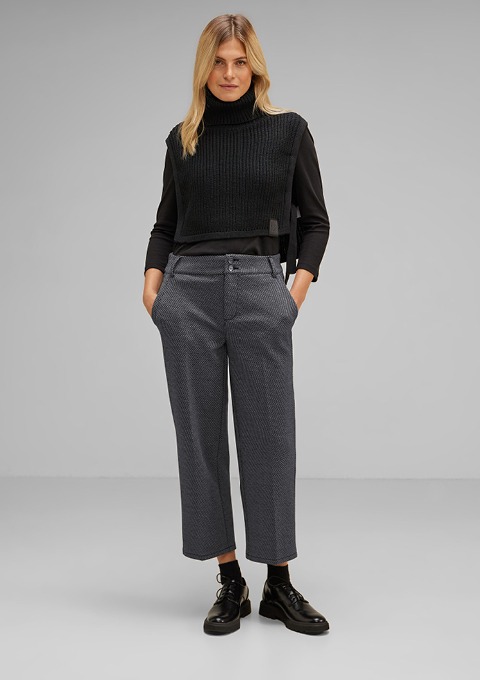 Eine Frau trägt einen schwarzen, kurzen Pullover und graue, gemusterte Hosen. Sie steht mit den Händen in den Taschen. Eine Frau trägt einen schwarzen, kurzen Pullover und graue, gemusterte Hosen. Sie steht mit den Händen in den Taschen.