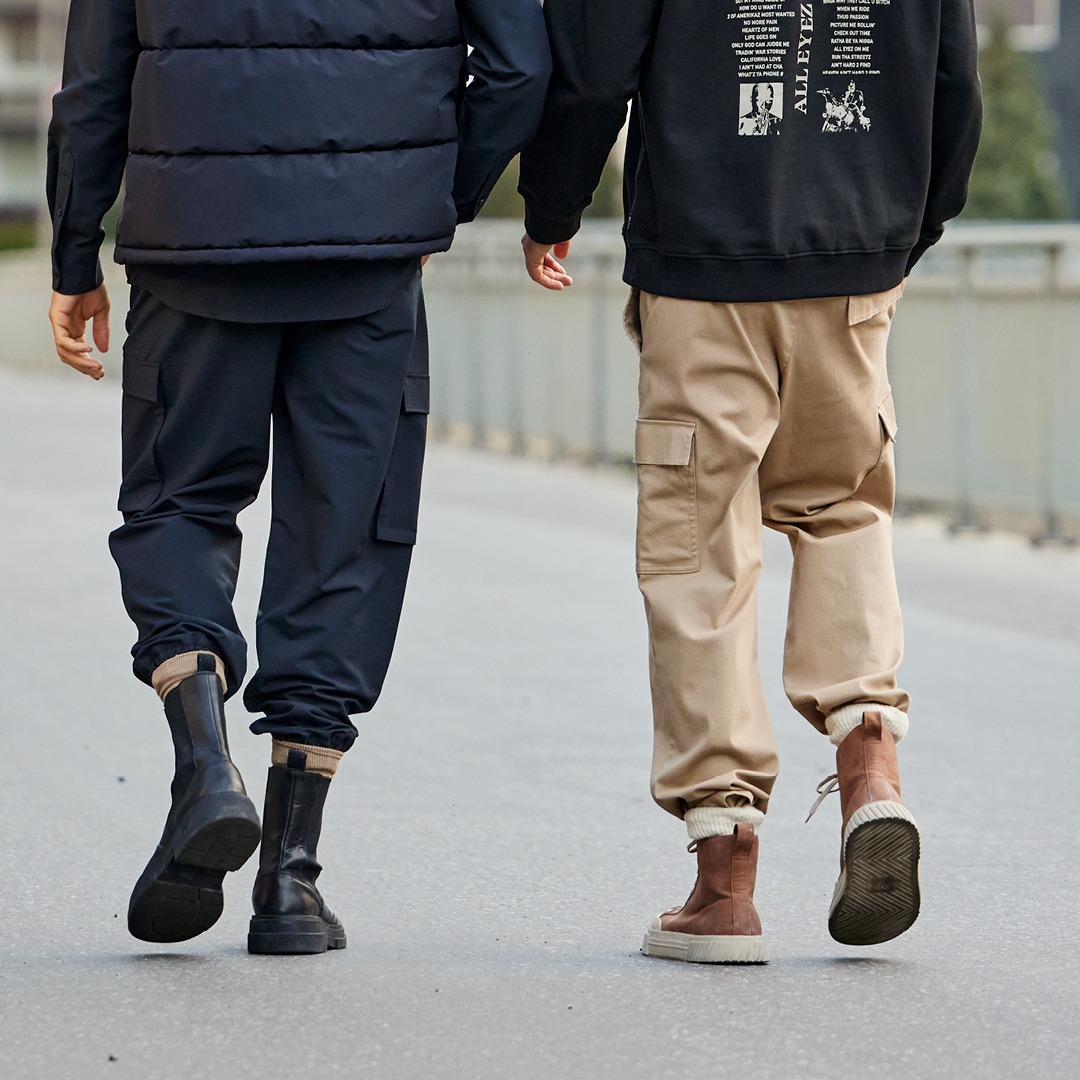 Zwei Personen gehen Hand in Hand auf einer Straße, eine trägt einen schwarzen Hoodie und beige Hosen, die andere ein dunkelblaues Outfit mit einer gesteppte Weste. Zwei Personen gehen Hand in Hand auf einer Straße, eine trägt einen schwarzen Hoodie und beige Hosen, die andere ein dunkelblaues Outfit mit einer gesteppte Weste.