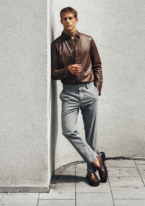 Ein Mann in einer braunen Lederjacke, grauen Hosen und braunen Schuhen lehnt an einer Wand. Ein Mann in einer braunen Lederjacke, grauen Hosen und braunen Schuhen lehnt an einer Wand.