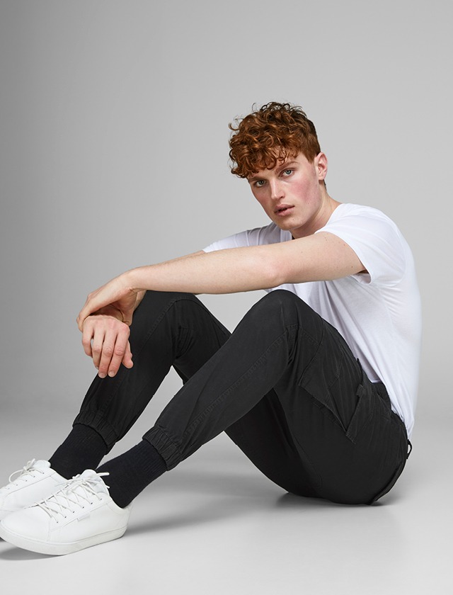 Ein junger Mann sitzt auf dem Boden, trägt ein weißes T-Shirt und schwarze Jogginghosen sowie weiße Sneakers. Ein junger Mann sitzt auf dem Boden, trägt ein weißes T-Shirt und schwarze Jogginghosen sowie weiße Sneakers.