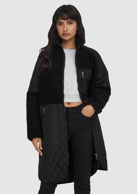 Eine Frau trägt einen schwarzen, gepolsterten Mantel mit Fleece- und Steppdesign, kombiniert mit einem grauen T-Shirt und schwarzen Hosen. Eine Frau trägt einen schwarzen, gepolsterten Mantel mit Fleece- und Steppdesign, kombiniert mit einem grauen T-Shirt und schwarzen Hosen.
