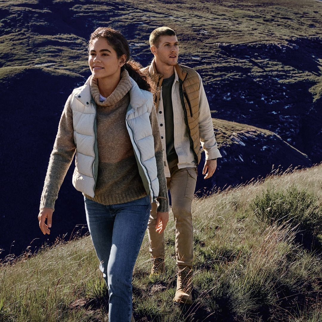Zwei Personen wandern auf einem Hügel, umgeben von grünen Wiesen und Bergen. Die Frau trägt eine helle Weste und einen Pulli, der Mann einen beigen Mantel.