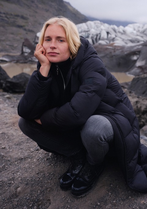 Eine Person in einer schwarzen Winterjacke sitzt im Freien, umgeben von einer felsigen Landschaft. Im Hintergrund sind Gletscher sichtbar. Eine Person in einer schwarzen Winterjacke sitzt im Freien, umgeben von einer felsigen Landschaft. Im Hintergrund sind Gletscher sichtbar.