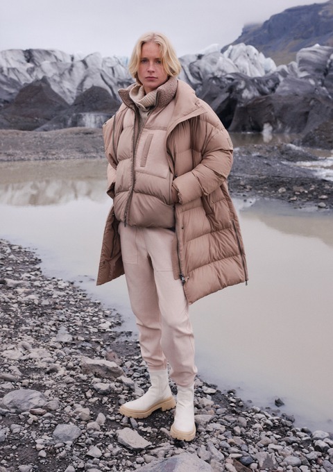 Eine Person steht an einem glacialen Landschaft mit einem beigen, gepolsterten Mantel und einer Hose. Eine Person steht an einem glacialen Landschaft mit einem beigen, gepolsterten Mantel und einer Hose.