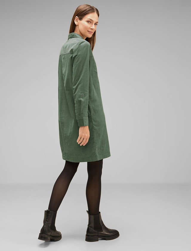 Eine Frau trägt ein grünes Hemdkleid und schwarze Stiefel, während sie von hinten zur Kamera blickt. Eine Frau trägt ein grünes Hemdkleid und schwarze Stiefel, während sie von hinten zur Kamera blickt.
