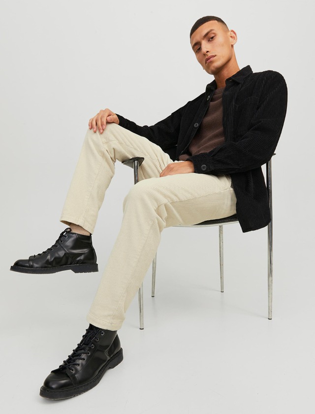 Ein junger Mann sitzt auf einem Stuhl, trägt ein schwarzes Hemd und hellbeige Hosen, sowie schwarze Stiefel. Ein junger Mann sitzt auf einem Stuhl, trägt ein schwarzes Hemd und hellbeige Hosen, sowie schwarze Stiefel.