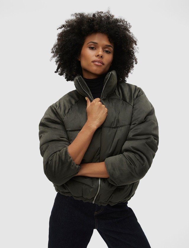 Eine Person mit lockigem Haar trägt eine grüne, kurzärmlige Jacke und posiert. Eine Person mit lockigem Haar trägt eine grüne, kurzärmlige Jacke und posiert.
