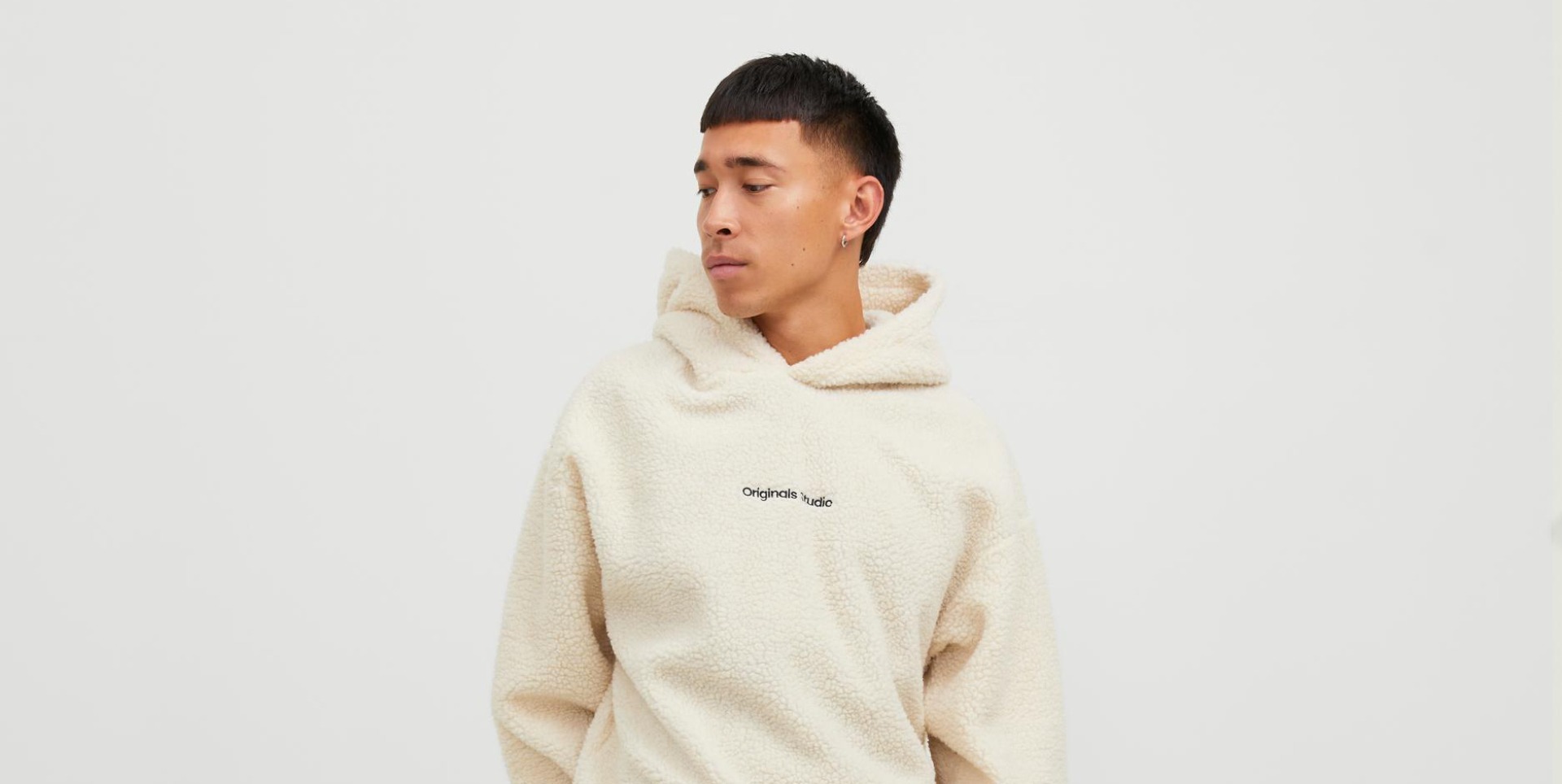 Ein junger Mann trägt einen beigen, flauschigen Hoodie mit der Aufschrift „Originals Studio“ auf der Brust.