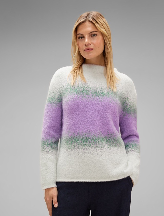 Eine Frau trägt einen pastellfarbenen Pullover mit lila und grünen Streifen vor einem grauen Hintergrund. Eine Frau trägt einen pastellfarbenen Pullover mit lila und grünen Streifen vor einem grauen Hintergrund.