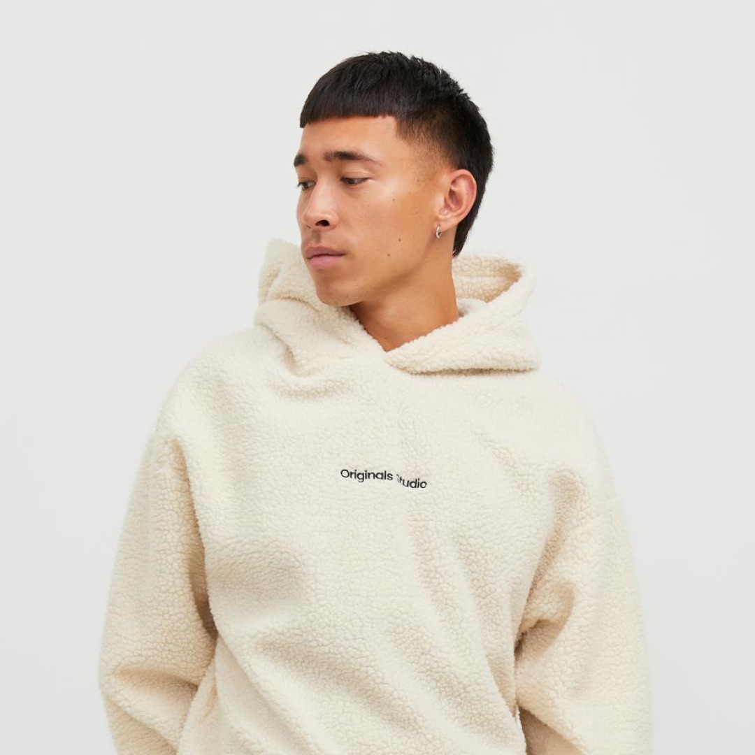 Ein Mann trägt einen cremefarbenen, flauschigen Hoodie mit der Aufschrift „Originals Studio“. Ein Mann trägt einen cremefarbenen, flauschigen Hoodie mit der Aufschrift "Originals Studio".