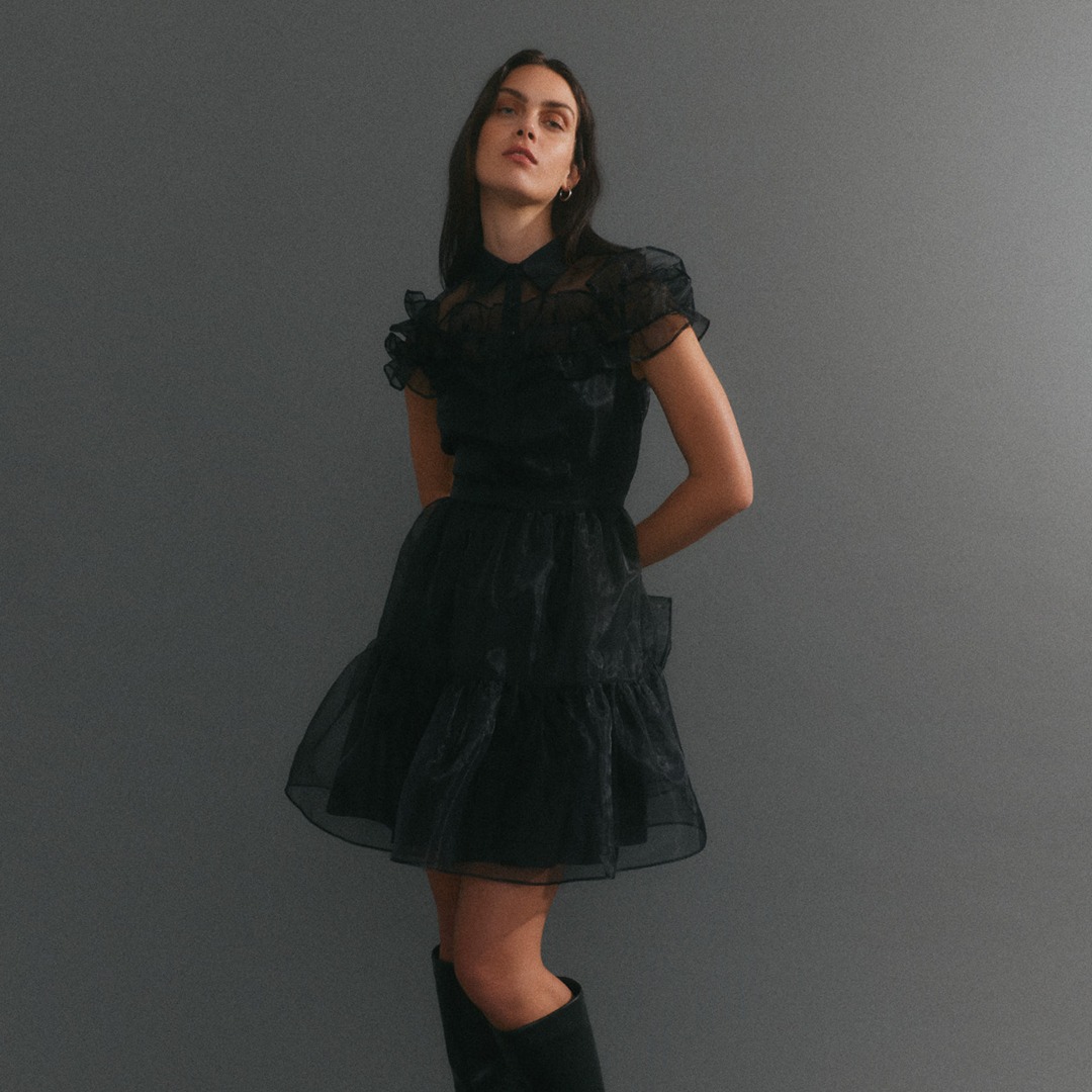 Eine Frau trägt ein schwarzes, volantes Kleid mit Rüschendetails vor einem grauen Hintergrund. Eine Frau trägt ein schwarzes, volantes Kleid mit Rüschendetails vor einem grauen Hintergrund.