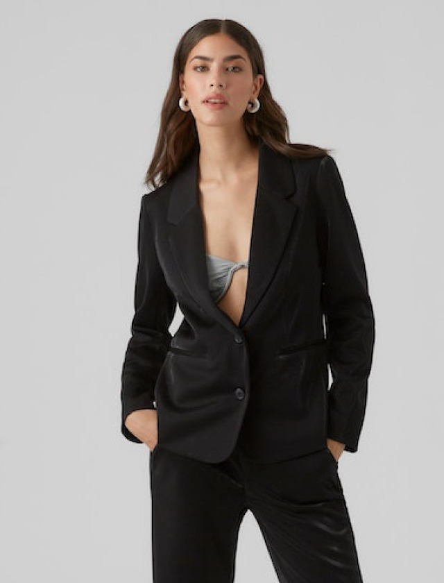 Eine Frau trägt einen schwarzen Blazer über einem grauen Oberteil. Sie hat lange, lockige Haare und große Ohrringe.