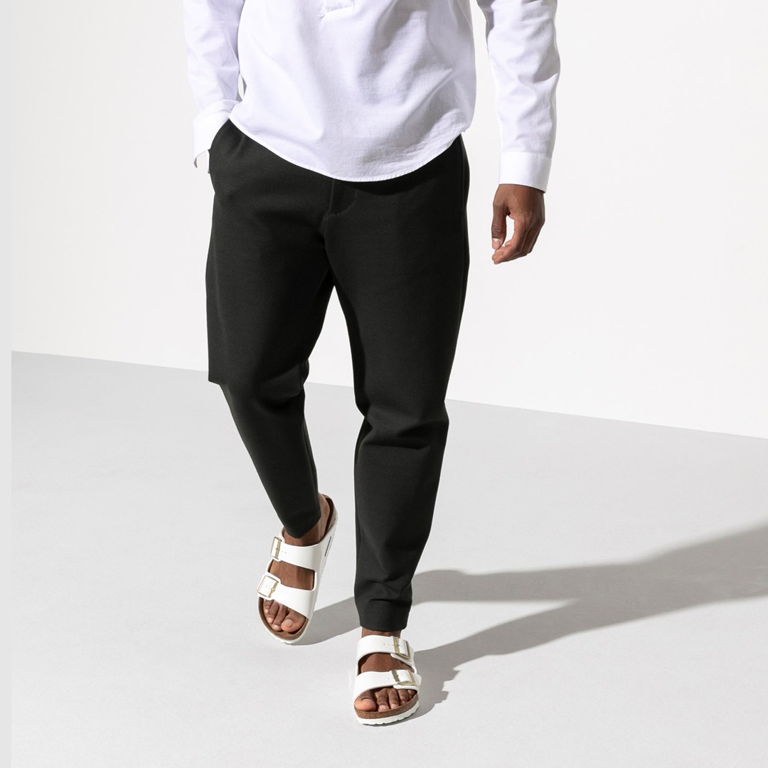 Eine Person trägt ein weißes Oberteil, schwarze Hose und weiße Sandalen.