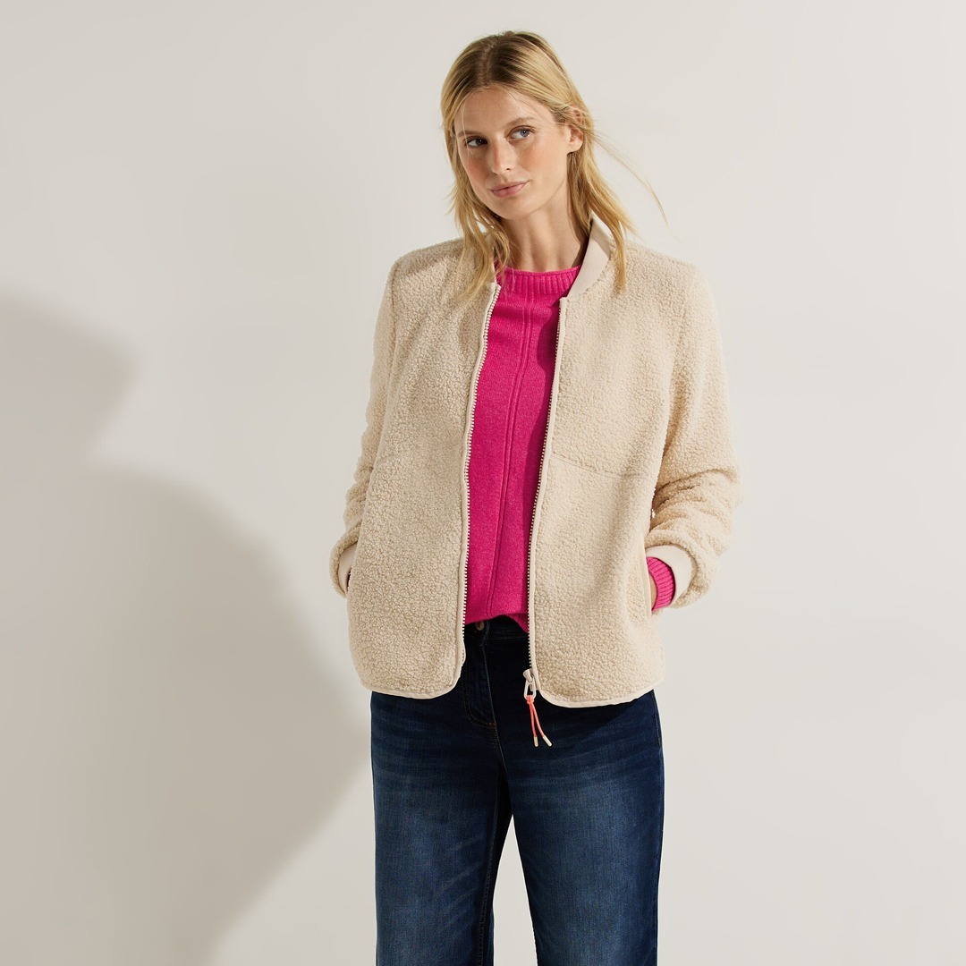 Eine Frau trägt einen beigen, flauschigen Mantel über einem pinken Pullover, kombiniert mit Jeans. Eine Frau trägt einen beigen, flauschigen Mantel über einem pinken Pullover, kombiniert mit Jeans.