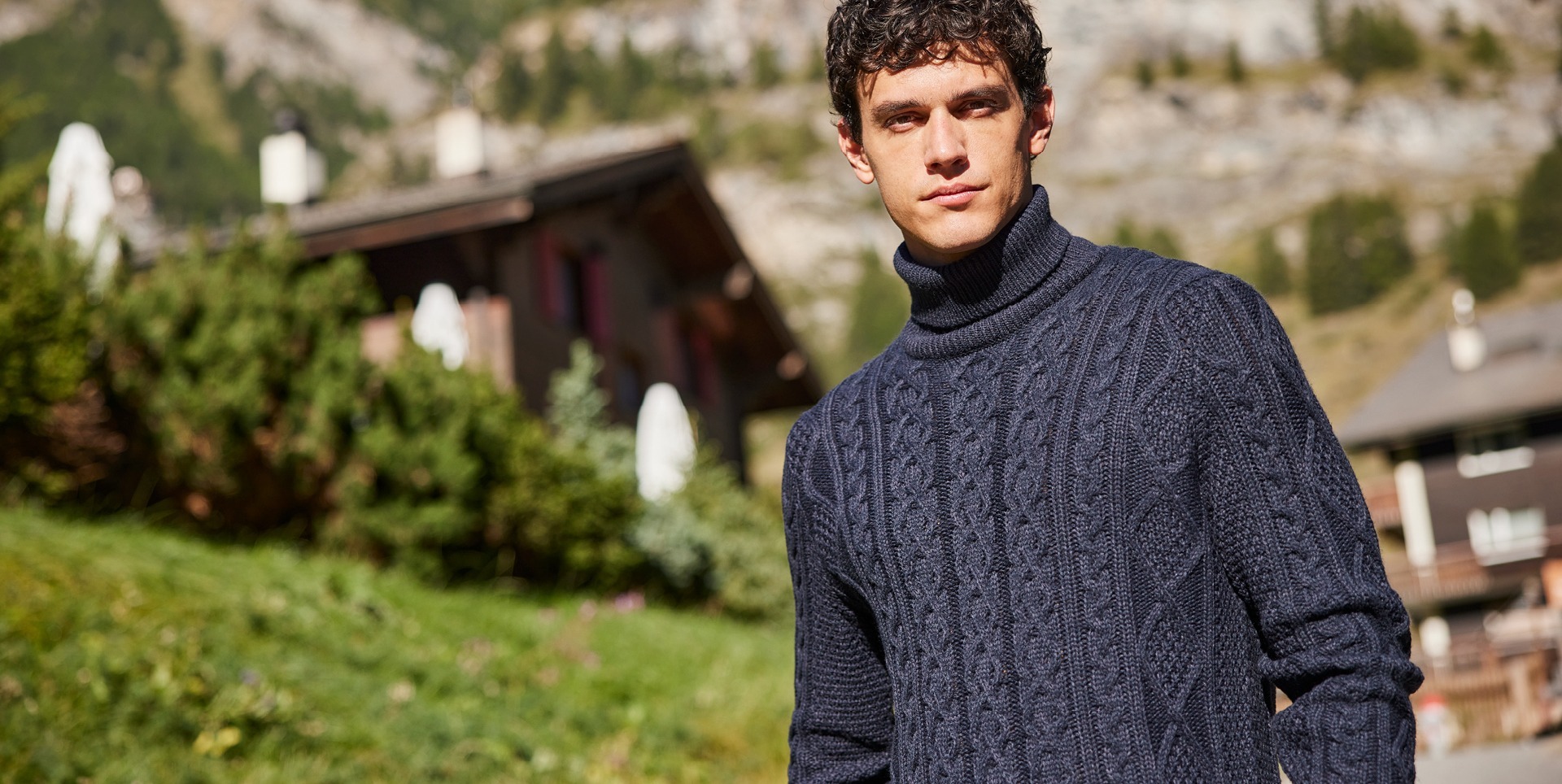 Ein Mann in einem blauen Rollkragenpullover steht im Freien, umgeben von Berglandschaft und Hütten. Ein Mann in einem blauen Rollkragenpullover steht im Freien, umgeben von Berglandschaft und Hütten.