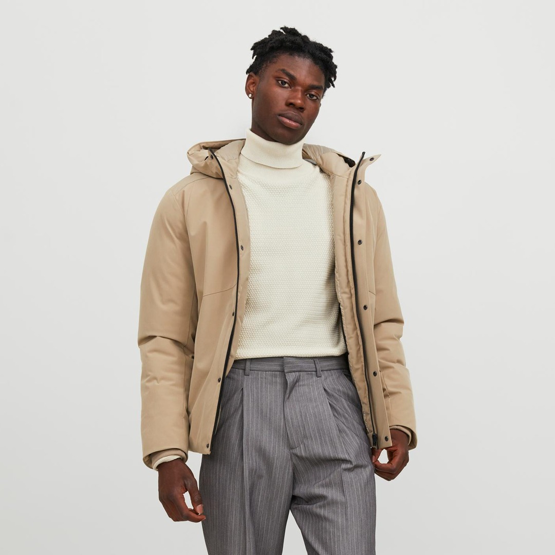 Ein junger Mann trägt eine beigefarbene Jacke und einen weißen Rollkragenpullover, kombiniert mit grauen Streifenhosen. Ein junger Mann trägt eine beigefarbene Jacke und einen weißen Rollkragenpullover, kombiniert mit grauen Streifenhosen.