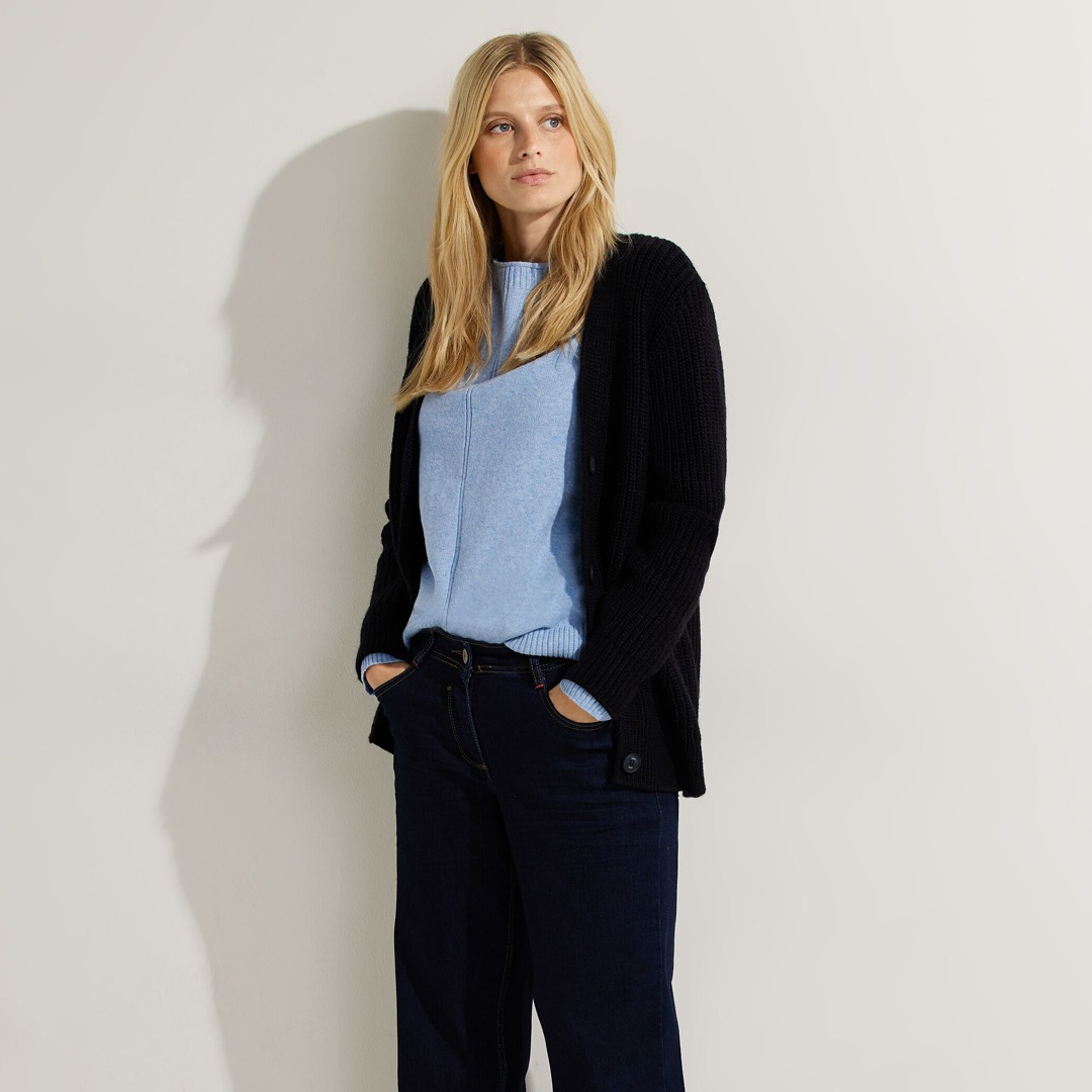Eine blonde Frau steht neben einer Wand, trägt einen blauen Pullover und eine schwarze Strickjacke sowie dunkelblaue Jeans. Eine blonde Frau steht neben einer Wand, trägt einen blauen Pullover und eine schwarze Strickjacke sowie dunkelblaue Jeans.