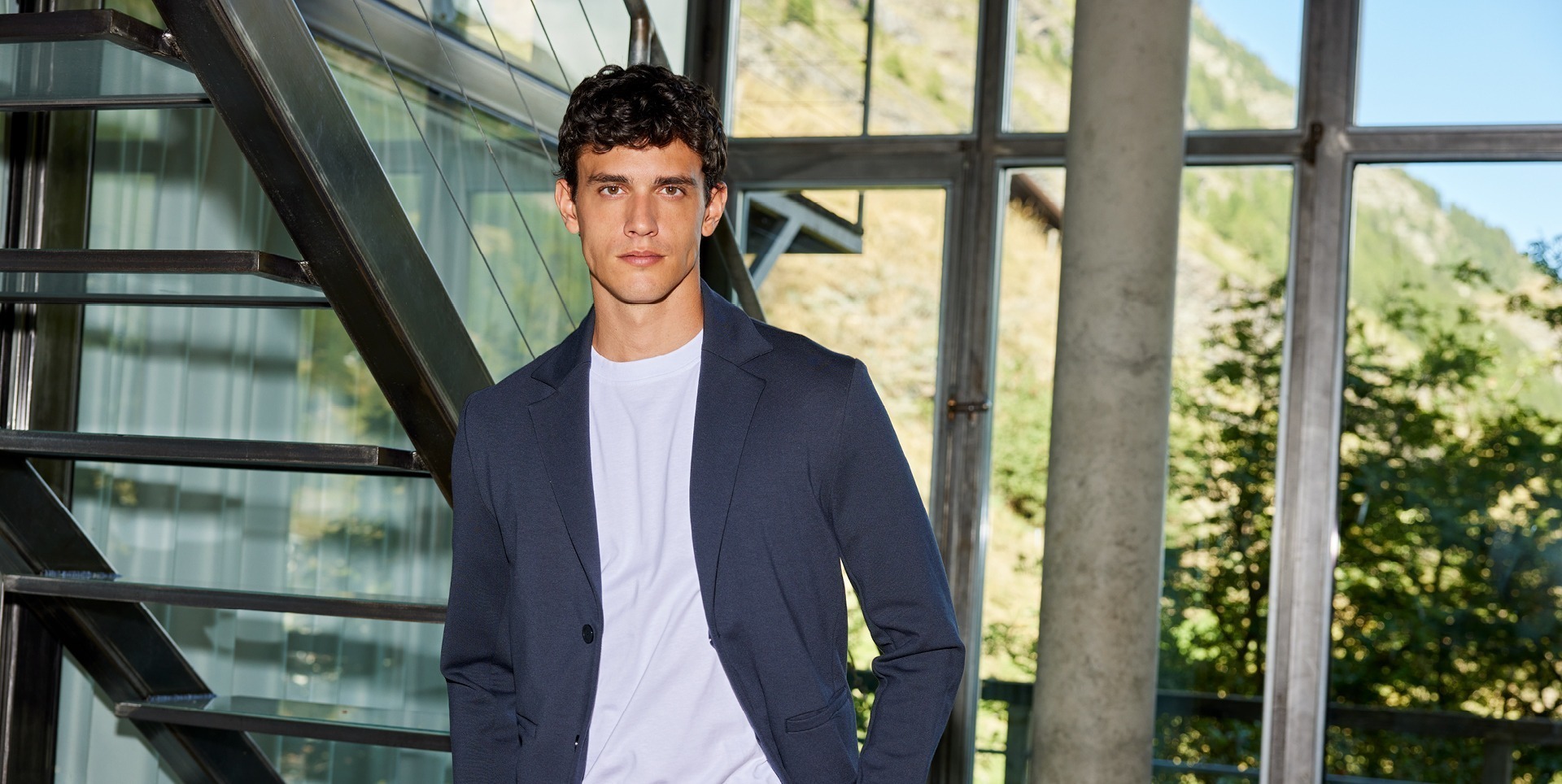 Ein junger Mann in einem dunklen Blazer und T-Shirt steht an einer Treppe in einem modernen Gebäude mit großen Fenstern und einer grünen Landschaft im Hintergrund. Ein junger Mann in einem dunklen Blazer und T-Shirt steht an einer Treppe in einem modernen Gebäude mit großen Fenstern und einer grünen Landschaft im Hintergrund.