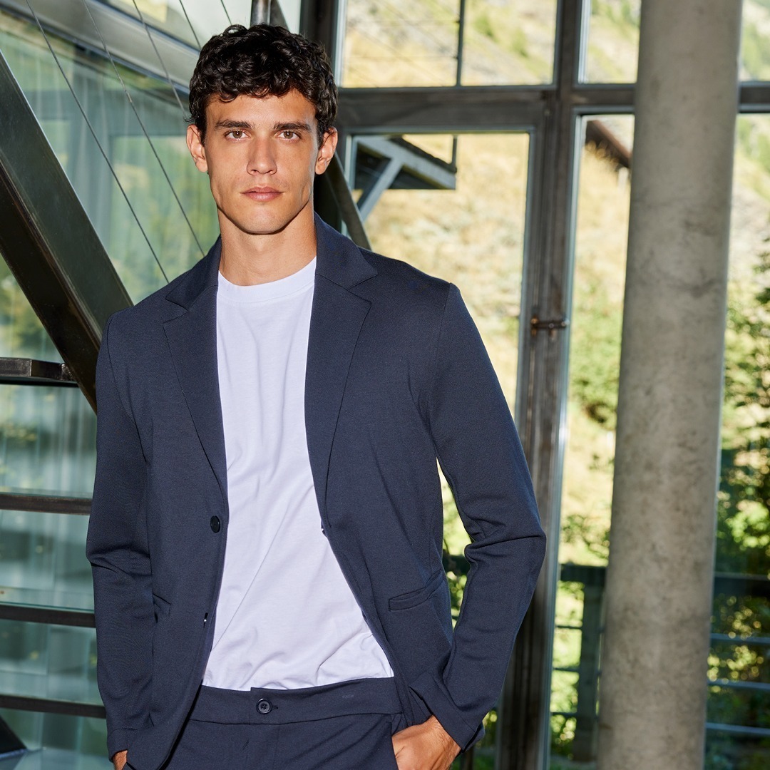 Ein junger Mann trägt einen dunkelblauen Blazer über einem weißen T-Shirt. Er steht an einer Treppe in einem modernen Gebäude. Ein junger Mann trägt einen dunkelblauen Blazer über einem weißen T-Shirt. Er steht an einer Treppe in einem modernen Gebäude.