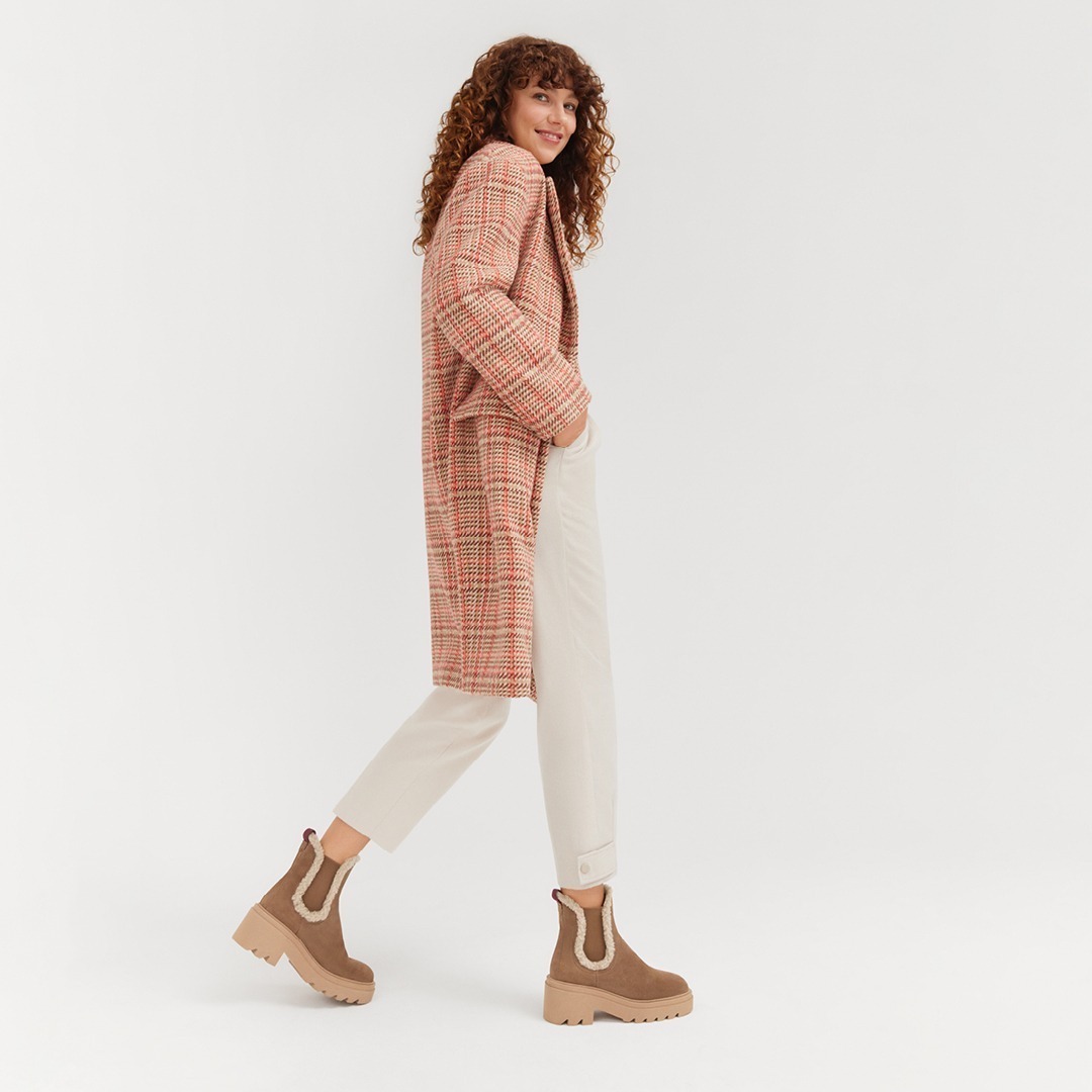 Eine lächelnde Frau mit lockigem Haar trägt einen karierten Mantel, beige Hosen und braune Stiefel.