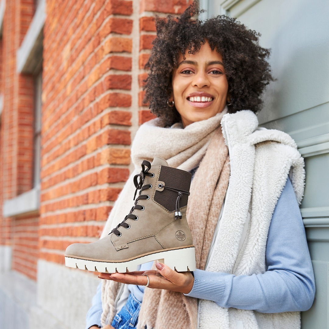 Eine Frau hält einen grauen Stiefel in der Hand und lächelt. Sie trägt einen Schal und eine warme Jacke.