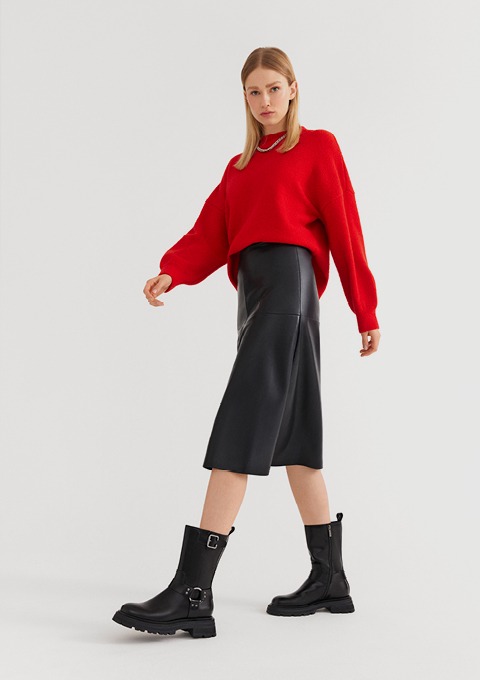 Ein Model trägt einen roten Pullover, einen schwarzen Midirock und schwarze Stiefel.