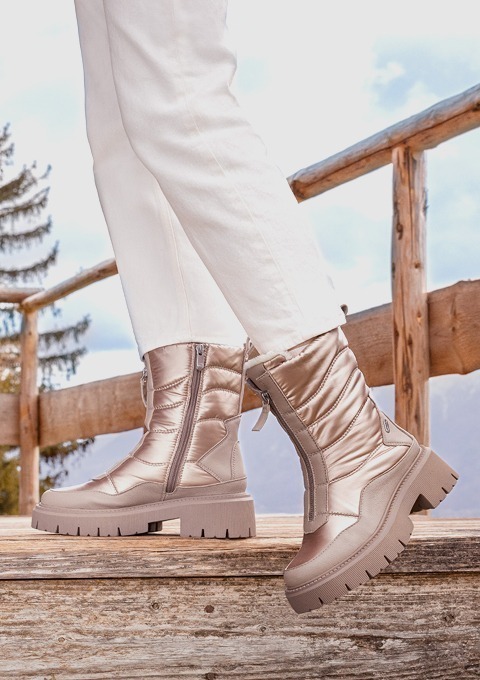 Schimmernde, beige Winterstiefel mit dickem Profil, getragen mit weißen Hosen, vor einer Holzsicht.