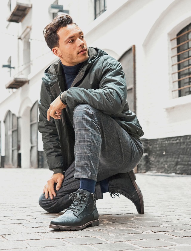 Ein Mann in einem grauen Parka und einer blauen Tasse kniet auf einem Steinboden und trägt eine graue Hose sowie gestiefelte Schuhe.