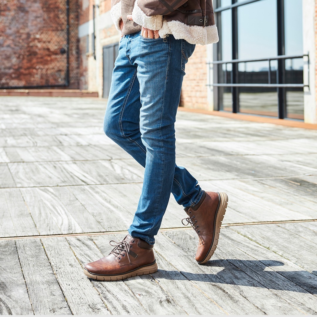 Eine Person trägt eine braune Jacke, einen beigen Pullover und blue Jeans, steht auf einem Holzboden und trägt braune Stiefel.