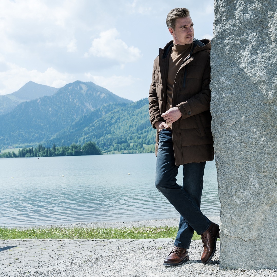 Ein Mann steht an einem See, umgeben von Bergen, trägt eine braune Jacke und Jeans.