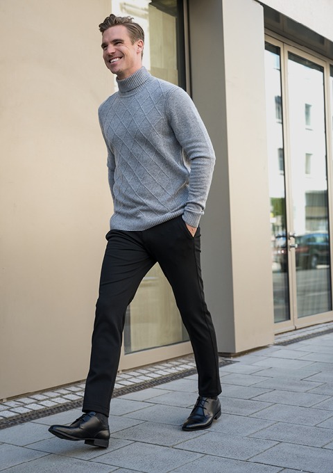 Ein lächelnder Mann trägt einen grauen Rollkragenpullover und schwarze Hosen, während er auf einem Gehweg entlanggeht.