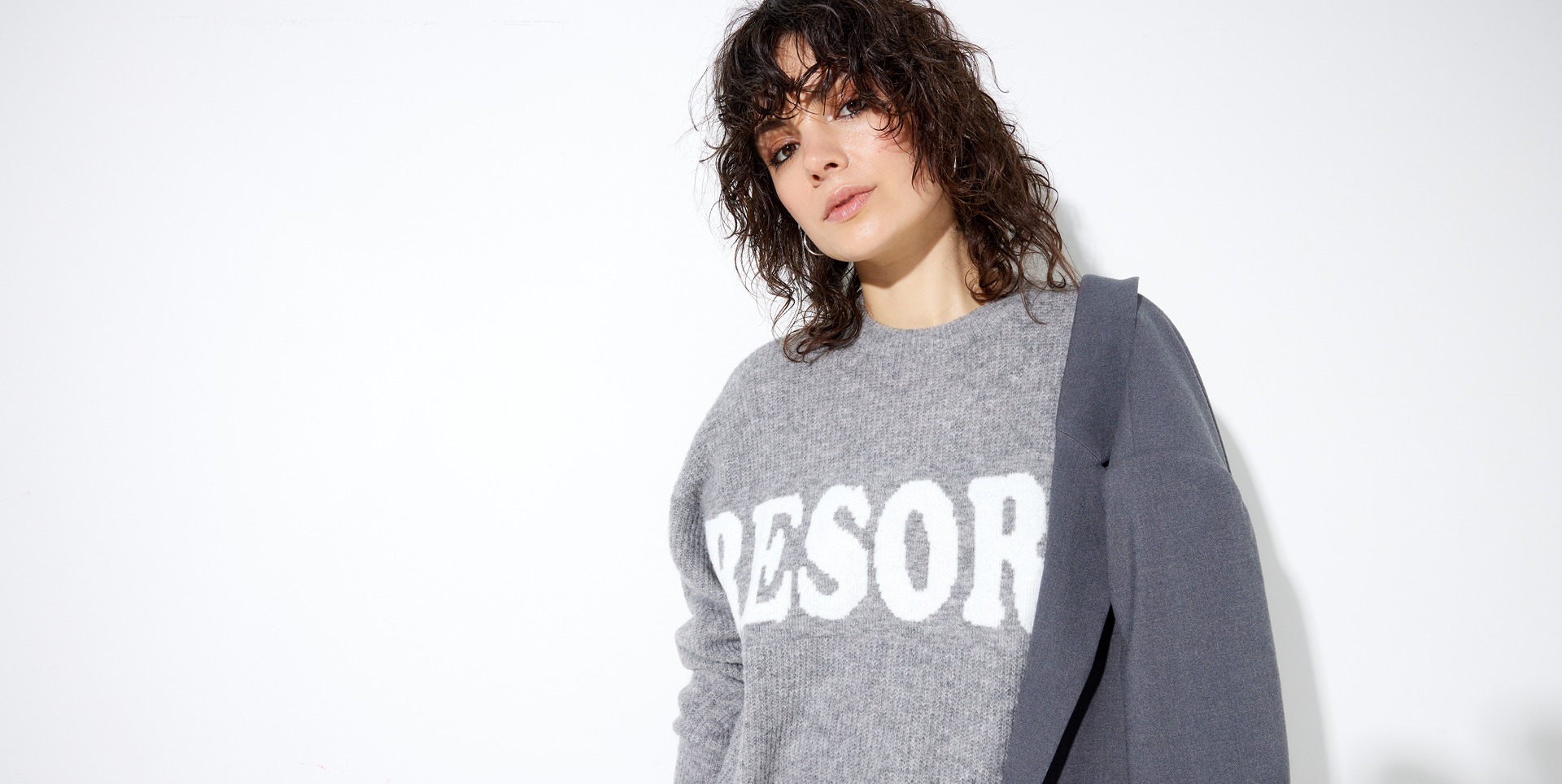 Eine Person mit lockigem Haar trägt einen grauen Pullover mit der Aufschrift "RESORT" und hat eine graue Jacke über die Schulter gelegt. Der Hintergrund ist weiß.