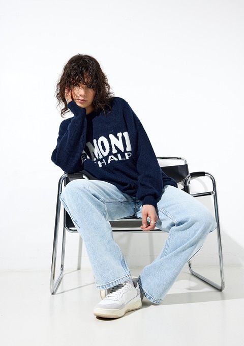 Eine Person mit lockigem Haar sitzt auf einem Stuhl. Sie trägt einen blauen Pullover mit Aufschrift und eine weite Jeans.