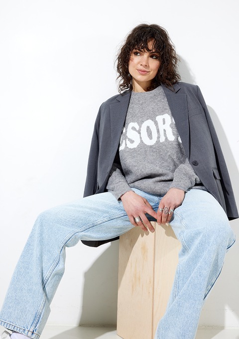 Eine Frau mit lockigem Haar sitzt auf einem Holzblock und trägt einen grauen Pullover mit Aufschrift sowie einen grauen Blazer. Sie hat eine entspannte Pose und lächelt.