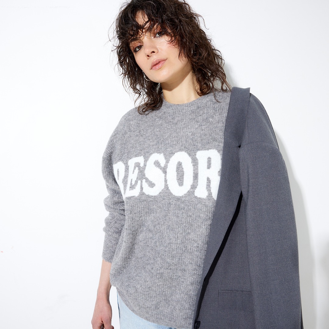 Eine Person trägt einen grauen Pullover mit der Aufschrift "RESORT" und hält einen grauen Blazer. Sie hat lockige Haare und schaut direkt in die Kamera.
