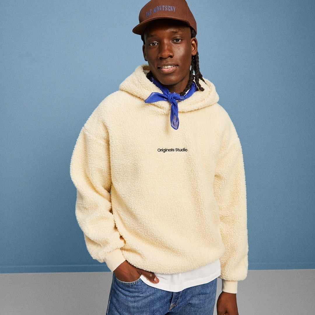 Ein junger Mann trägt einen beigefarbenen Hoodie mit Kapuze und einer blauen Halstuch. Er steht vor einer blauen Wand.