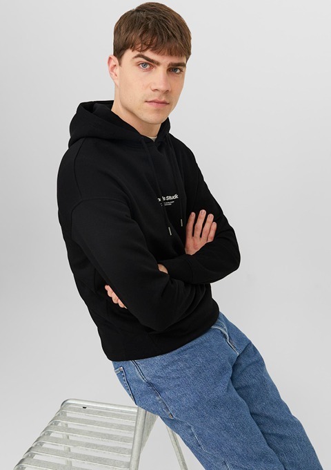 Ein junger Mann sitzt auf einer Leiter und trägt einen schwarzen Hoodie sowie jeans.