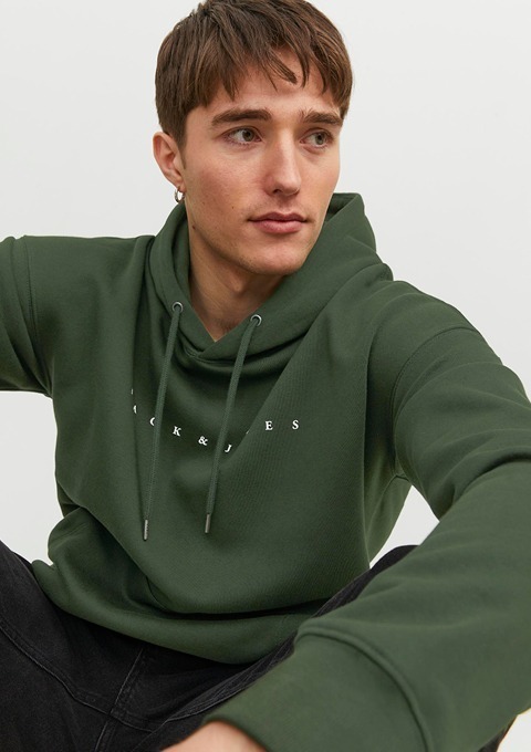 Ein junger Mann trägt einen grünen Hoodie und sitzt entspannt mit einem neutralen Ausdruck.