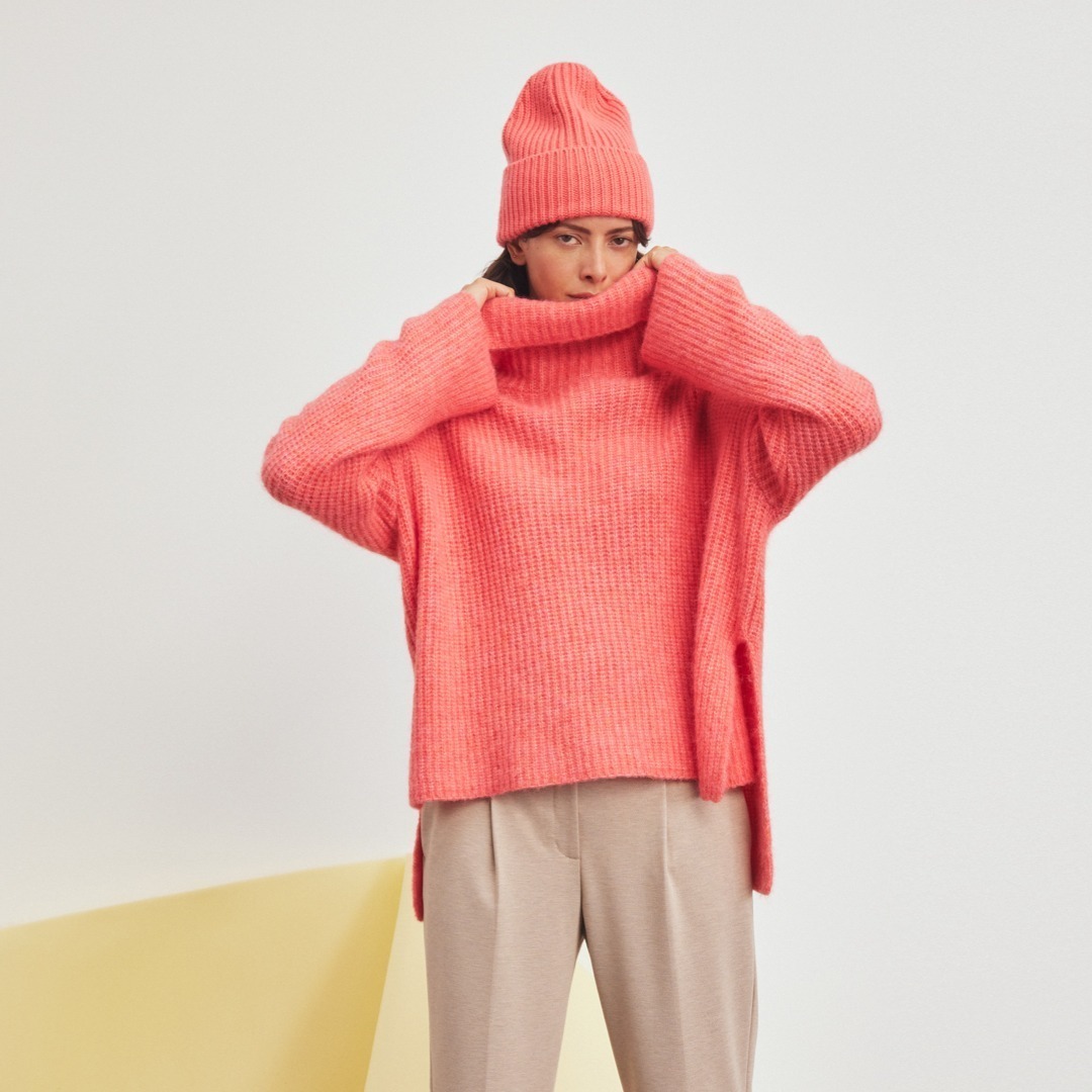 Eine Person trägt einen pinken Pullover und eine Mütze, sie zieht den Pullover hoch. Der Hintergrund ist neutral.
