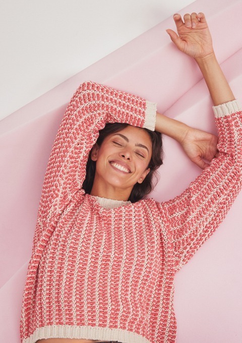 Eine lächelnde Frau trägt einen rosa Pullover und breitet ihre Arme aus, vor einem rosa Hintergrund.