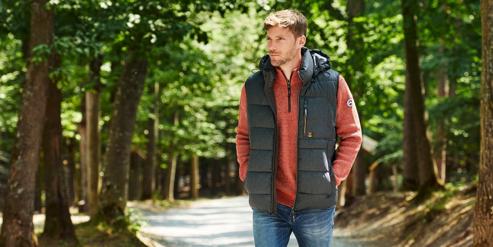 Ein Mann in einer grauen Weste und einem roten Pullover geht auf einem Waldweg. Bäume umgeben den Weg. Ein Mann in einer grauen Weste und einem roten Pullover geht auf einem Waldweg. Bäume umgeben den Weg.