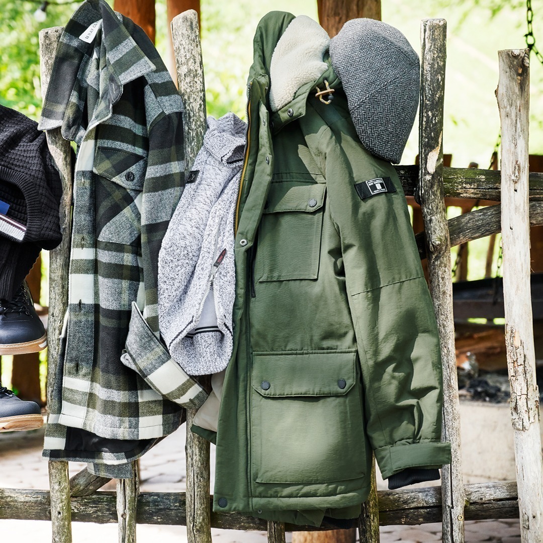 Kleidung hängt an einer Holzwand: eine grüne Jacke, ein grauer Pullover und ein kariertes Hemd. Kleidung hängt an einer Holzwand: eine grüne Jacke, ein grauer Pullover und ein kariertes Hemd.