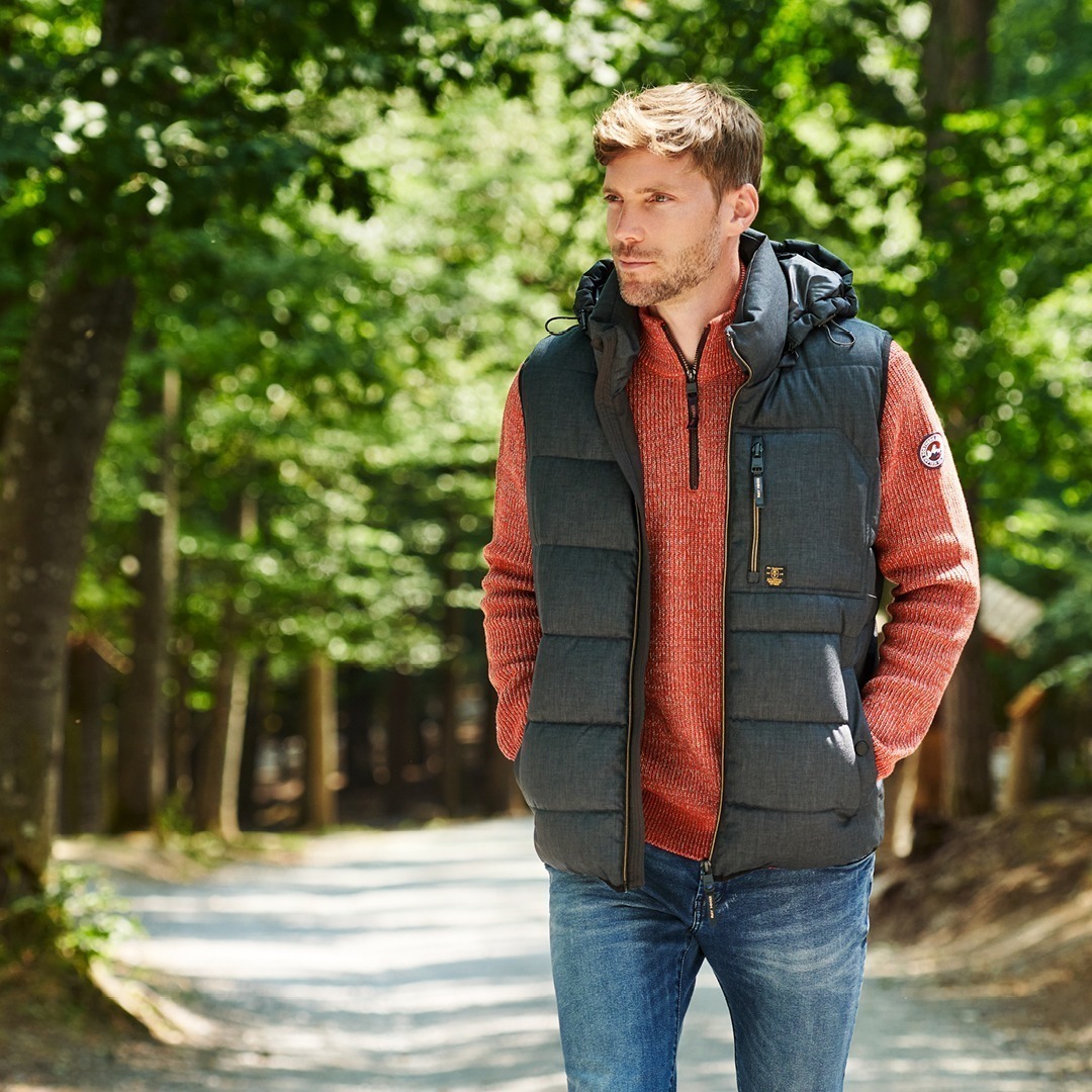 Ein Mann in einem grünen Steppwesten und einem roten Pullover steht in einem Wald auf einem Weg.
