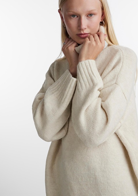 Eine Person trägt einen beigen Pullover und hat die Hände am Hals. Eine Person trägt einen beigen Pullover und hat die Hände am Hals.