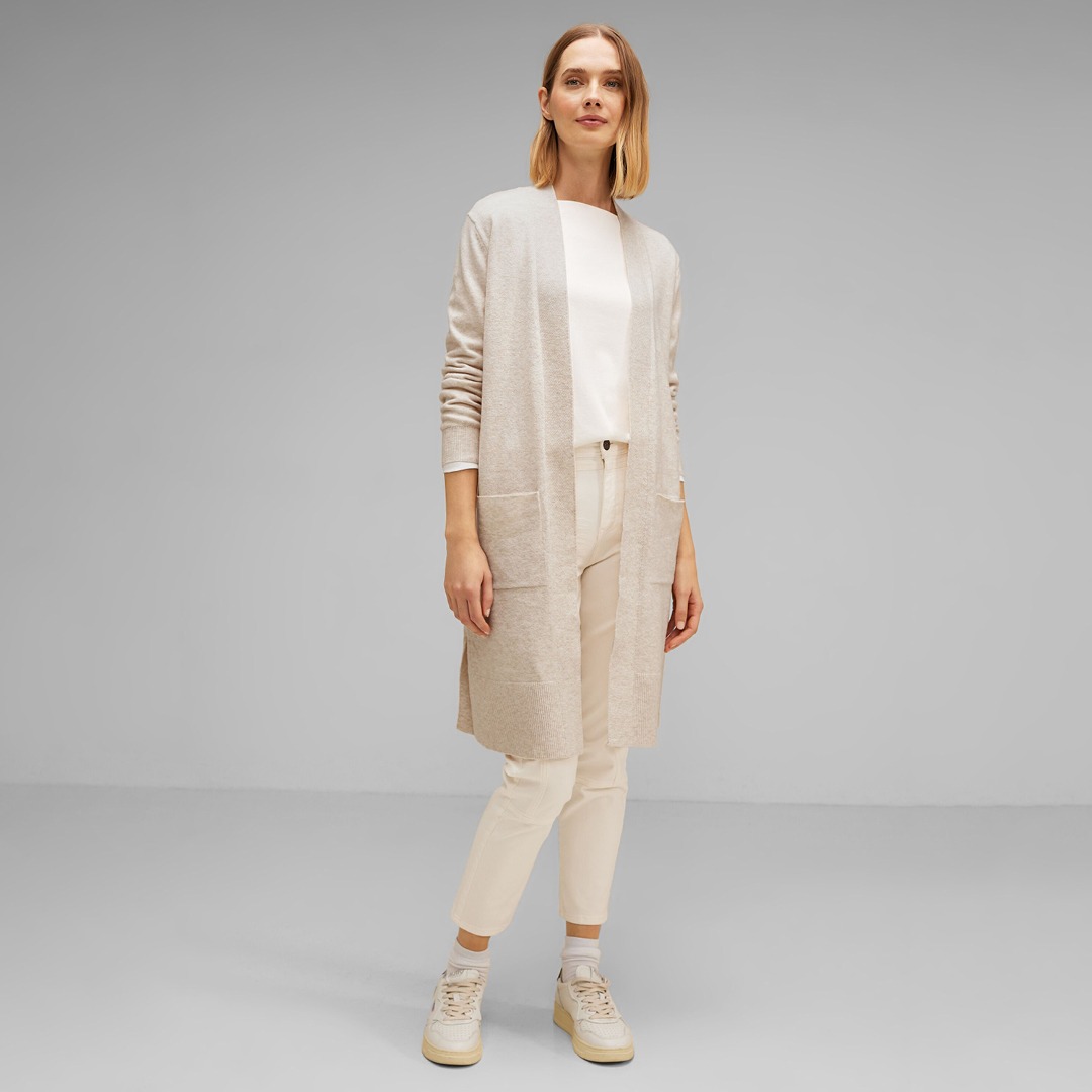 Eine Person steht in einem hellen, eleganten Outfit mit einem langen Cardigan, einem weißen Shirt und hellen Hosen. Eine Person steht in einem hellen, eleganten Outfit mit einem langen Cardigan, einem weißen Shirt und hellen Hosen.